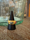 Gold ring with a red gemstone on a black cone-shaped stand in front of a green glass jar.