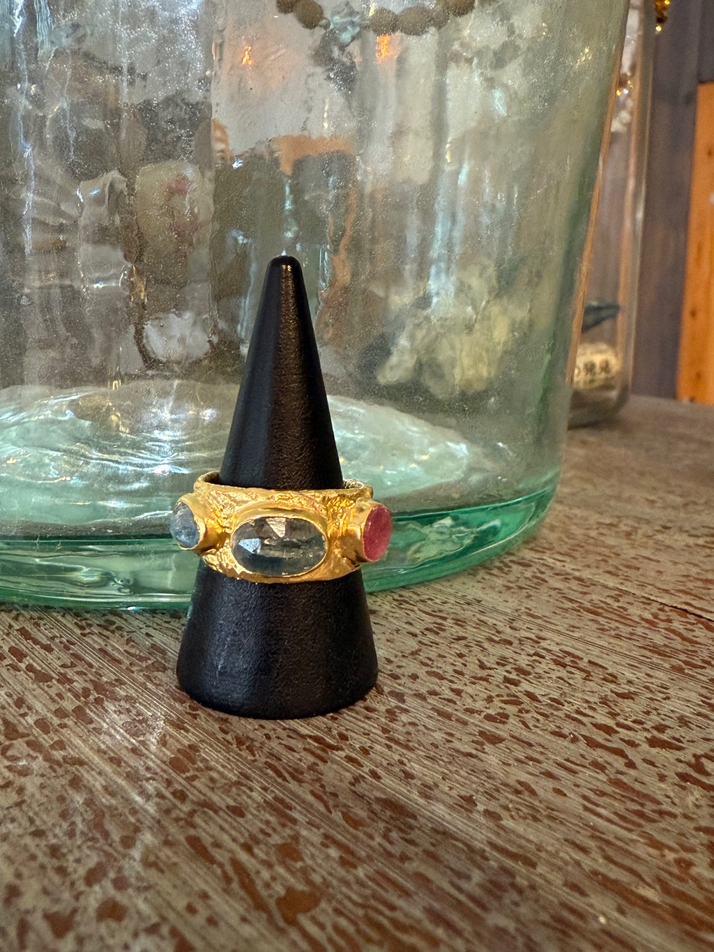 Gold ring with a red gemstone on a black cone-shaped stand in front of a green glass jar.