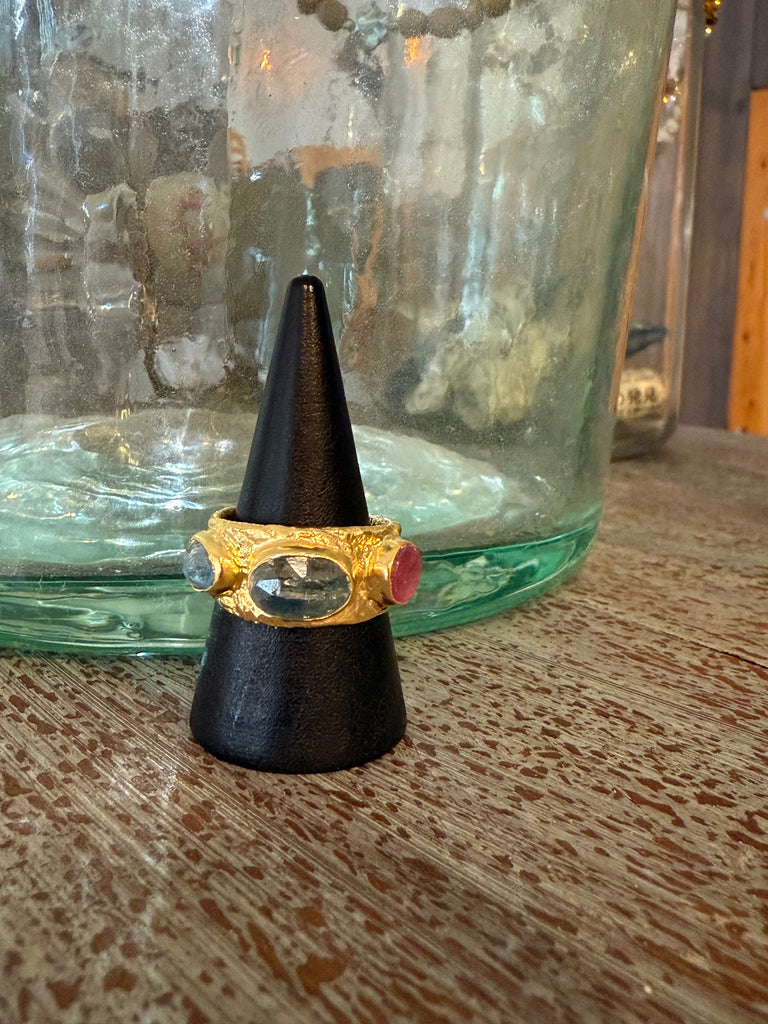 Gold ring with a red gemstone on a black cone-shaped stand in front of a green glass jar.