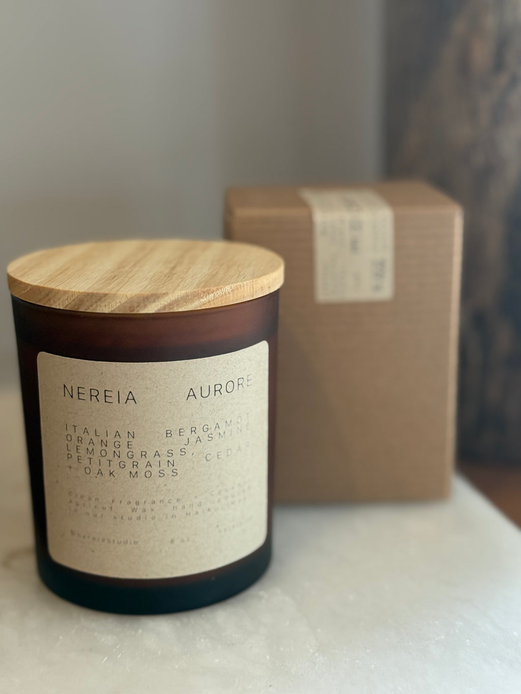 Candle labeled 'Nereia Aurore' with a wooden lid on a white surface, with a brown box in the background.