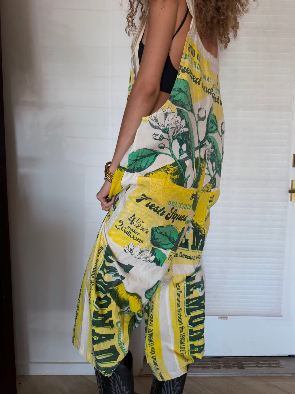 A woman wearing a sleeveless wide-leg jumpsuit with a vintage lemonade print featuring yellow lemons, green leaves, and white background with black text, styled with a black tank top and black boots.
