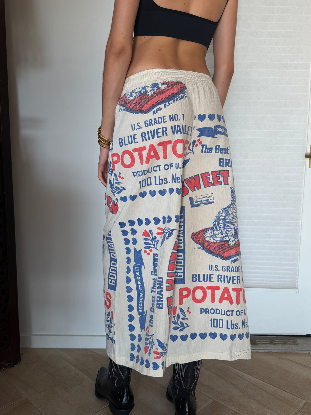 A person wearing wide-leg cream drawstring pants with a vintage-style red and blue potato print, paired with a black crop top and black boots, standing indoors against a white paneled wall.