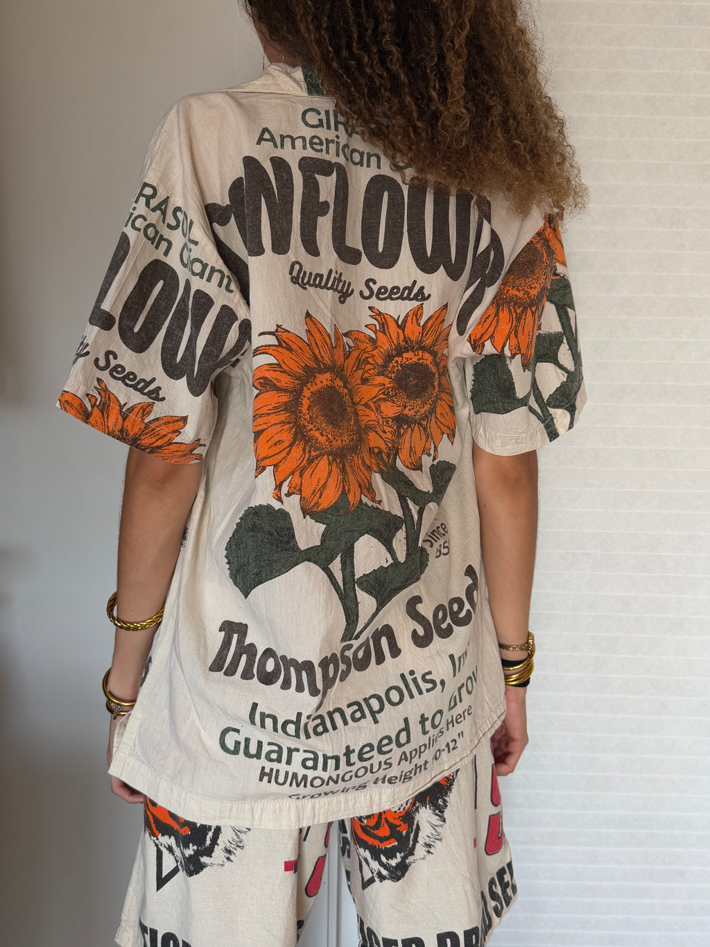 A woman wearing a short-sleeve button-front cover-up dress with a vintage-style sunflower and text print. The garment has an off-white base with large orange sunflowers, green leaves, and black typography, including 'Simpson' and 'Flowers'.