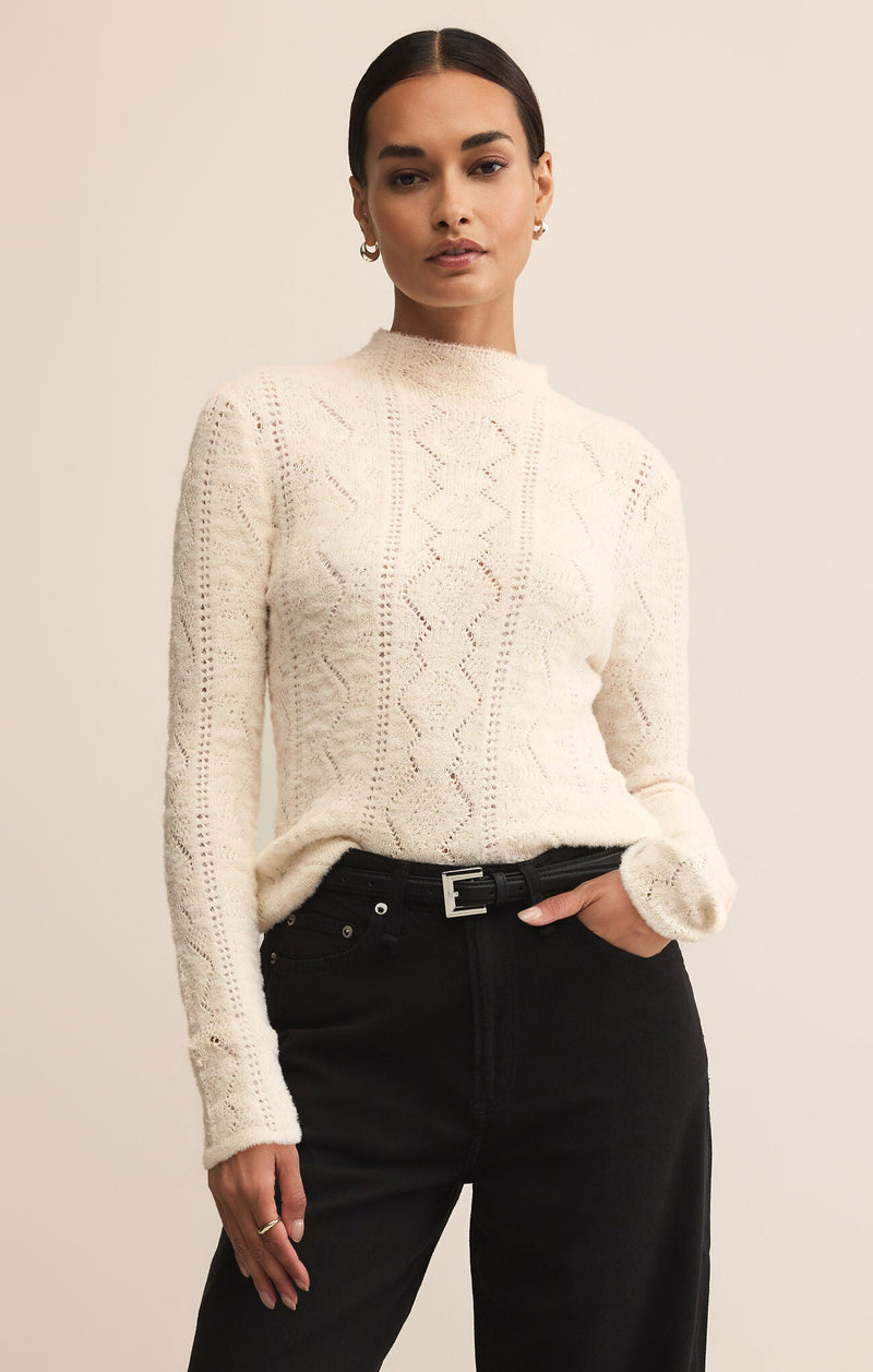 Woman wearing a cream cable knit sweater and black pants against a beige background