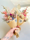 Bouquet of dried flowers wrapped in brown paper with a visible brand label, held by a person against a light background.
